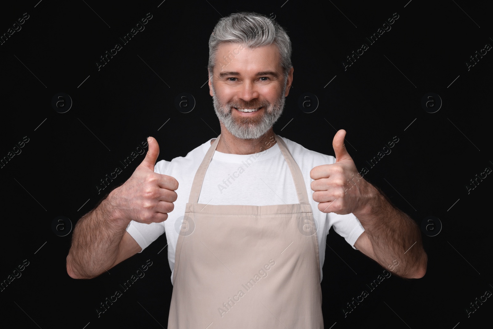 Photo of Happy man in kitchen apron showing thumbs up on black background. Mockup for design