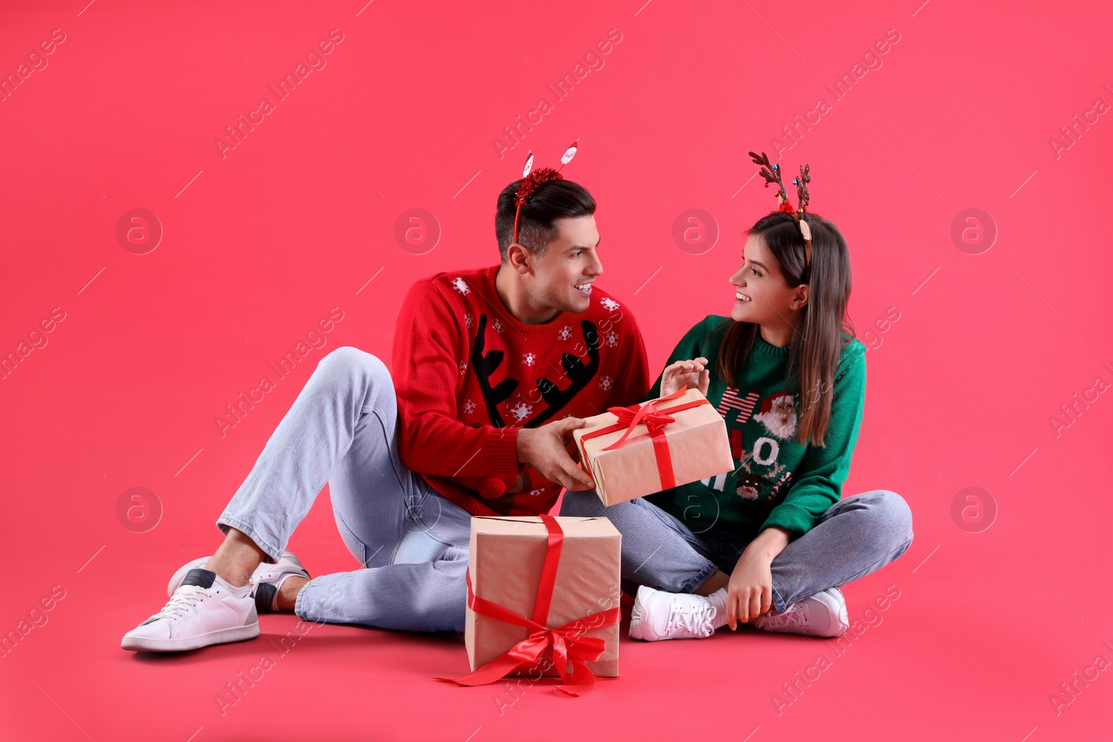 Photo of Beautiful happy couple in Christmas headbands and sweaters sitting with gifts on red background