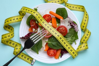 Photo of Measuring tape, vegetable salad and fork on light blue background, flat lay. Weight loss concept