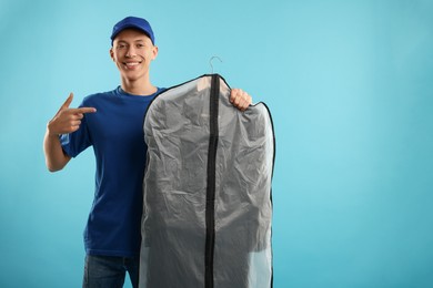 Dry-cleaning delivery. Happy courier holding garment cover with clothes on light blue background, space for text