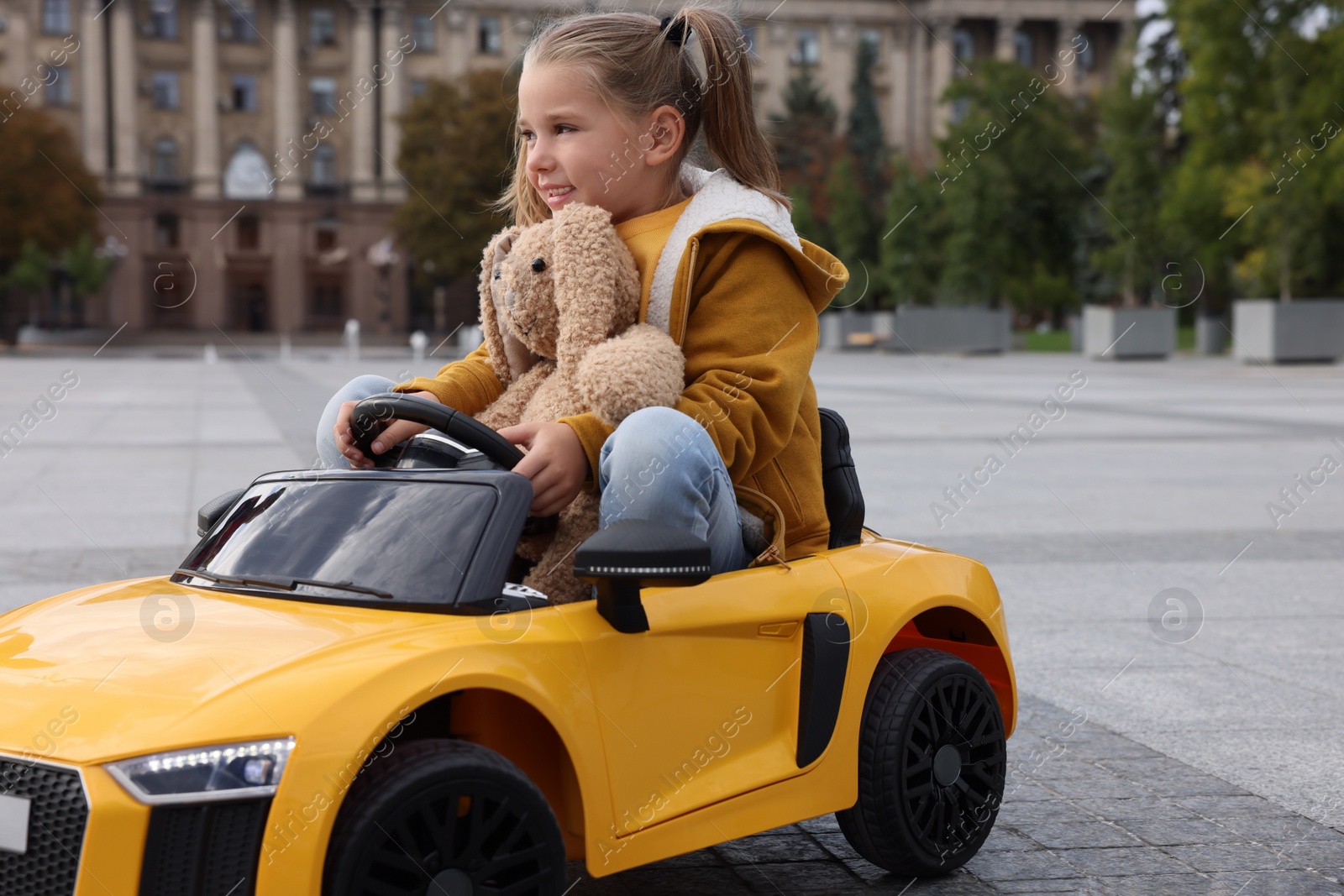 Photo of Cute little girl with toy bunny driving children's car on city street