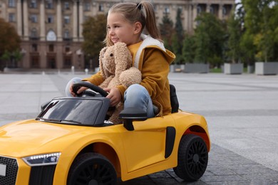 Cute little girl with toy bunny driving children's car on city street