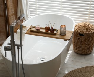 Photo of Wooden tray with cosmetic products, burning candles, and reed air freshener on bath tub in bathroom