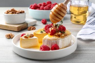 Photo of Pouring honey onto brie cheese served with raspberries and walnuts on white wooden table