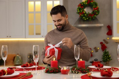 Happy young man opening Christmas gift at table in kitchen