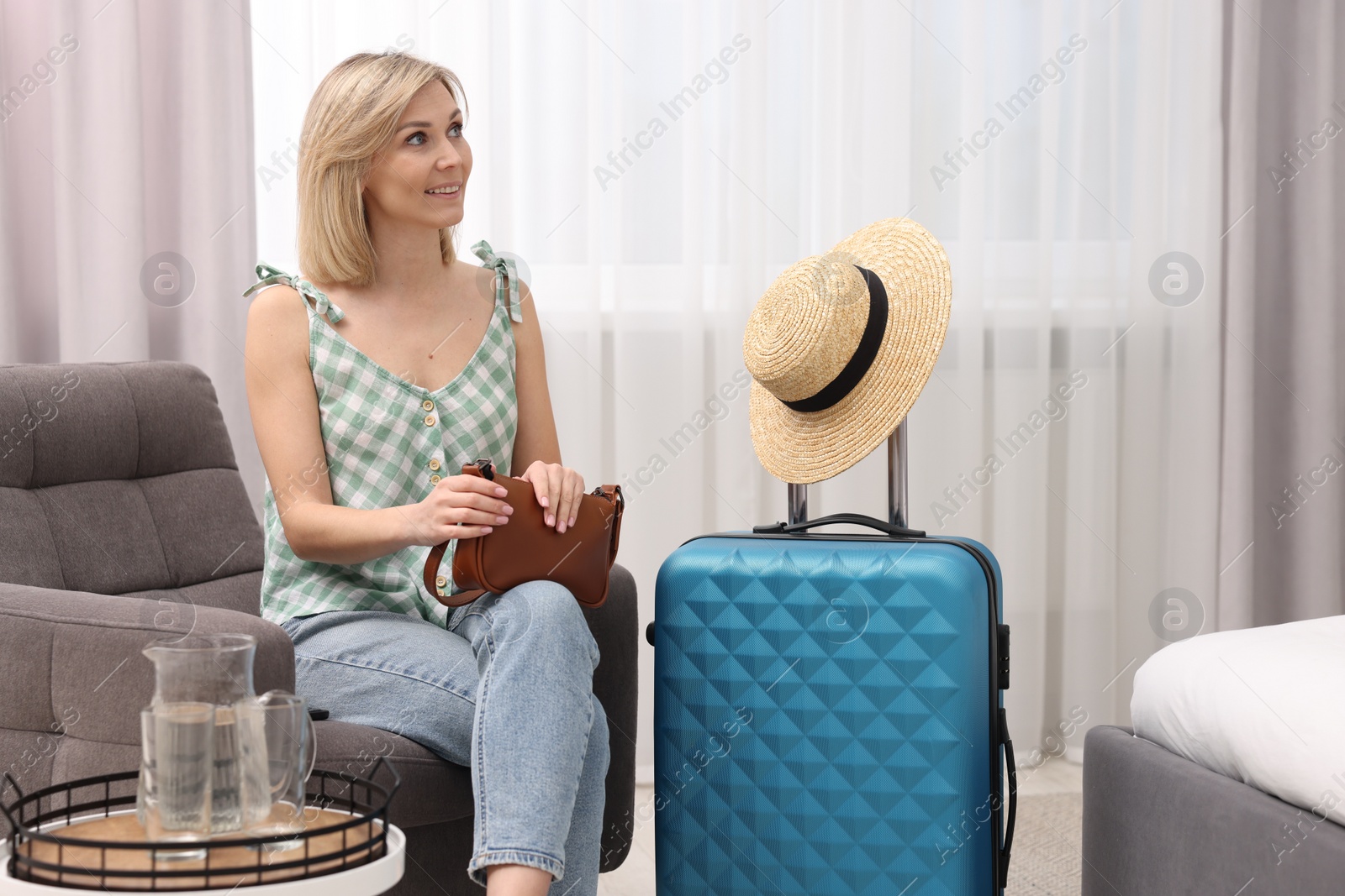 Photo of Smiling guest with bag relaxing on armchair in stylish hotel room