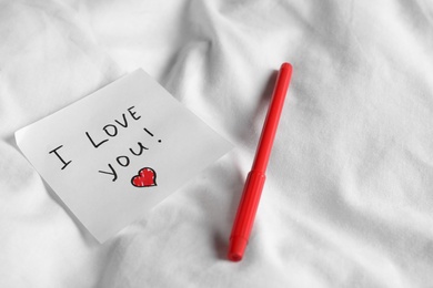 Photo of Sticky note with phrase I love you! and red felt pen on bed, space for text