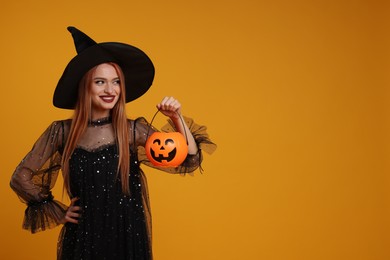Photo of Happy young woman in scary witch costume with pumpkin bucket on orange background, space for text. Halloween celebration
