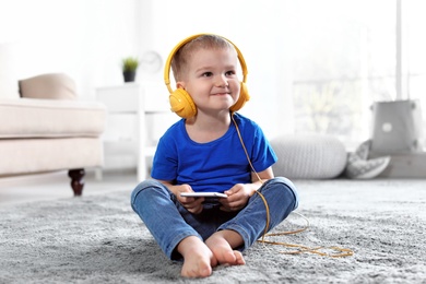 Photo of Cute child with headphones and mobile phone on floor indoors