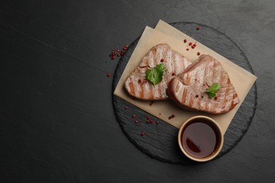 Delicious tuna steaks with sauce, parsley and spices on black table, top view. Space for text