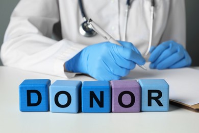 Word Donor made of wooden cubes. Doctor writing in notebook at white table, closeup