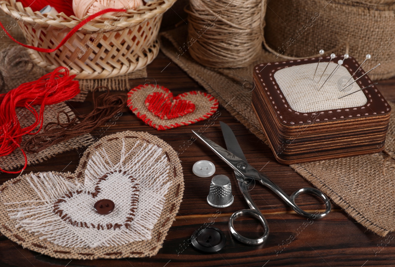 Photo of Heart shaped pieces of burlap fabric with different stitches and sewing tools on wooden table, closeup