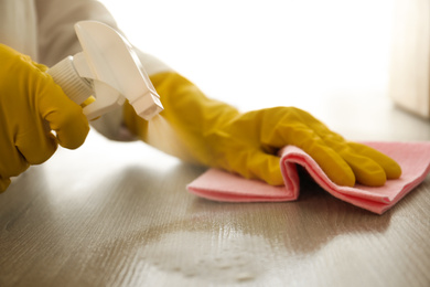 Photo of Woman in gloves cleaning wooden table with spray detergent and rag indoors, closeup