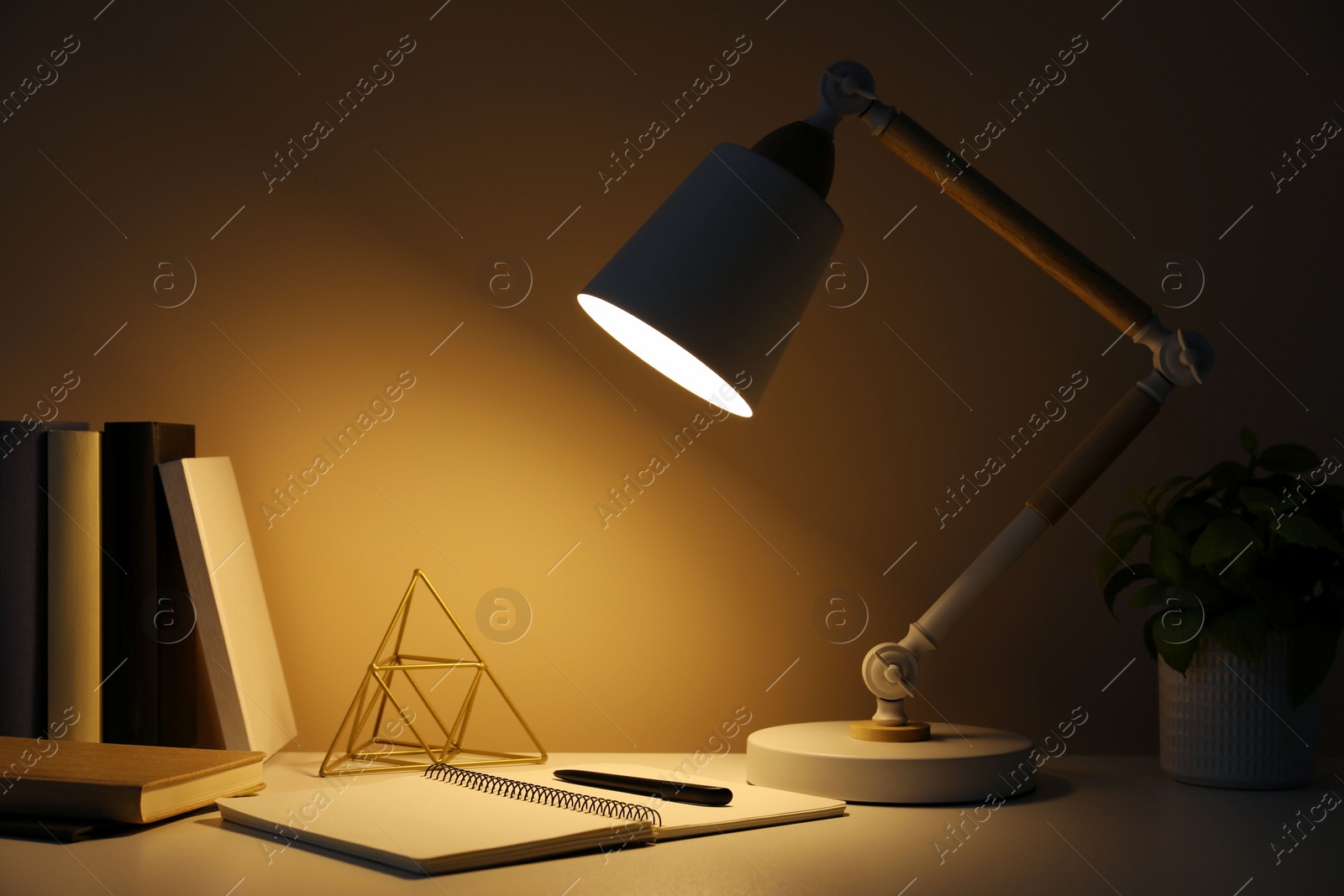 Photo of Stylish modern desk lamp, open notebook, books and plant on table near wall in dark room