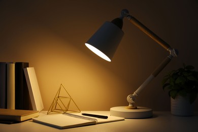 Photo of Stylish modern desk lamp, open notebook, books and plant on table near wall in dark room