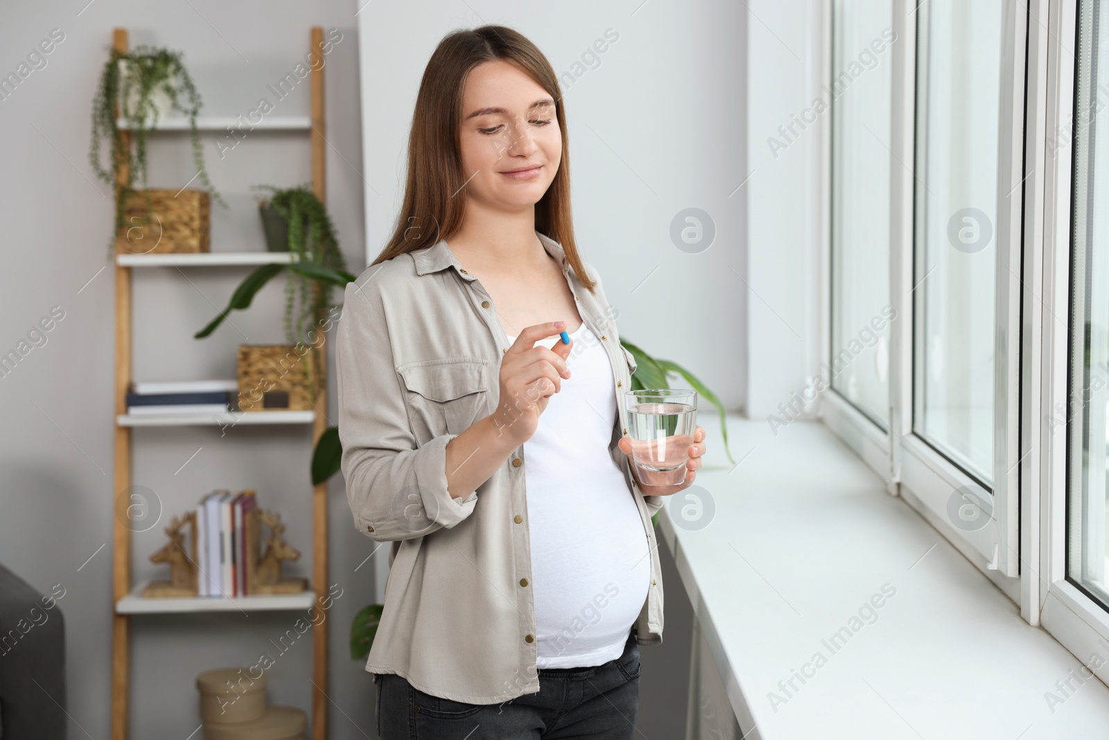 Photo of Beautiful pregnant woman holding pill and glass of water near window at home