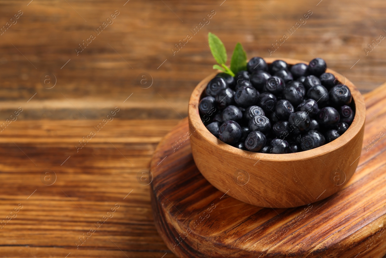 Photo of Ripe bilberries and leaves in bowl on wooden table, closeup. Space for text