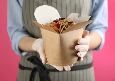 Photo of Chef holding box of wok noodles with seafood on pink background, closeup
