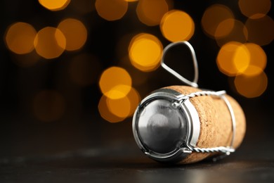 Photo of Sparkling wine cork with muselet cap on black table against blurred festive lights, closeup. Space for text