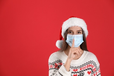Photo of Pretty woman in Santa hat and medical mask showing silence gesture on red background, space for text