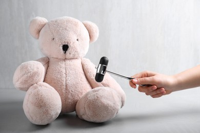 Photo of Woman pretending to test teddy bear's reflexes with hammer on grey background, closeup. Nervous system diagnostic
