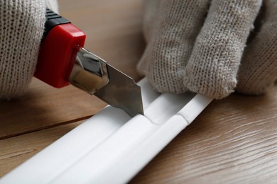 Photo of Worker cutting foam crown molding with utility knife at wooden table, closeup