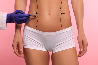 Image of Woman preparing for cosmetic surgery, pink background. Doctor drawing markings on her abdomen, closeup