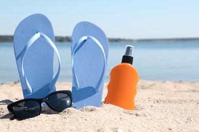 Photo of Stylish flip flops, sunglasses and sun protection spray on sandy beach