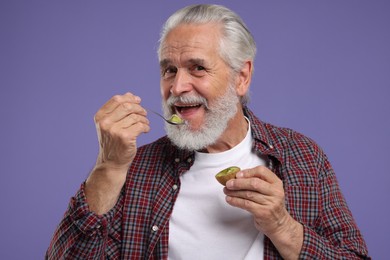Happy senior man eating kiwi with spoon on purple background