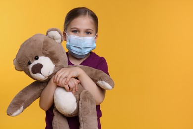 Cute girl with medical mask and teddy bear on orange background, space for text