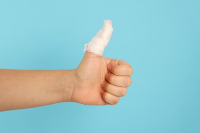 Photo of Woman showing thumb up wrapped in medical bandage on light blue background, closeup