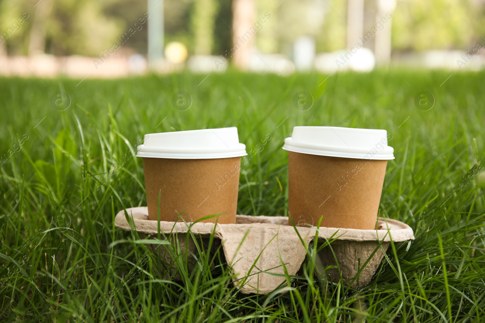 Photo of Takeaway paper coffee cups with plastic lids in cardboard holder on green grass outdoors