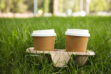 Takeaway paper coffee cups with plastic lids in cardboard holder on green grass outdoors