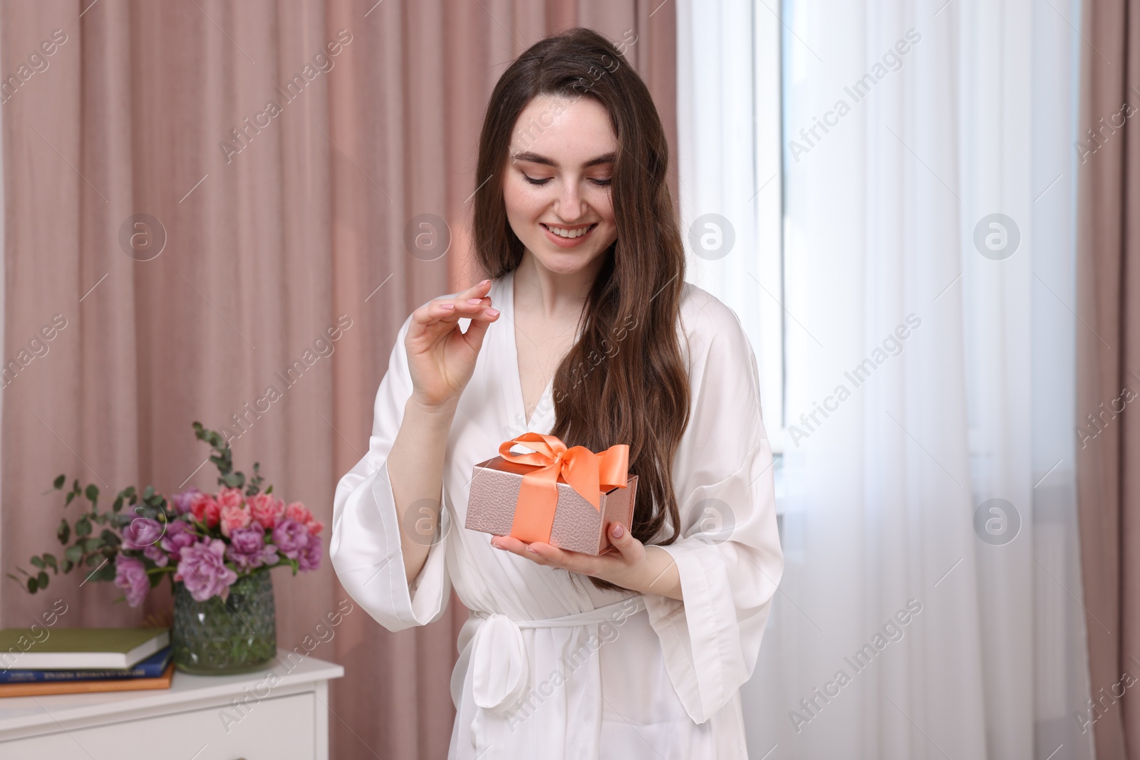 Photo of Beautiful happy young woman holding gift box in room