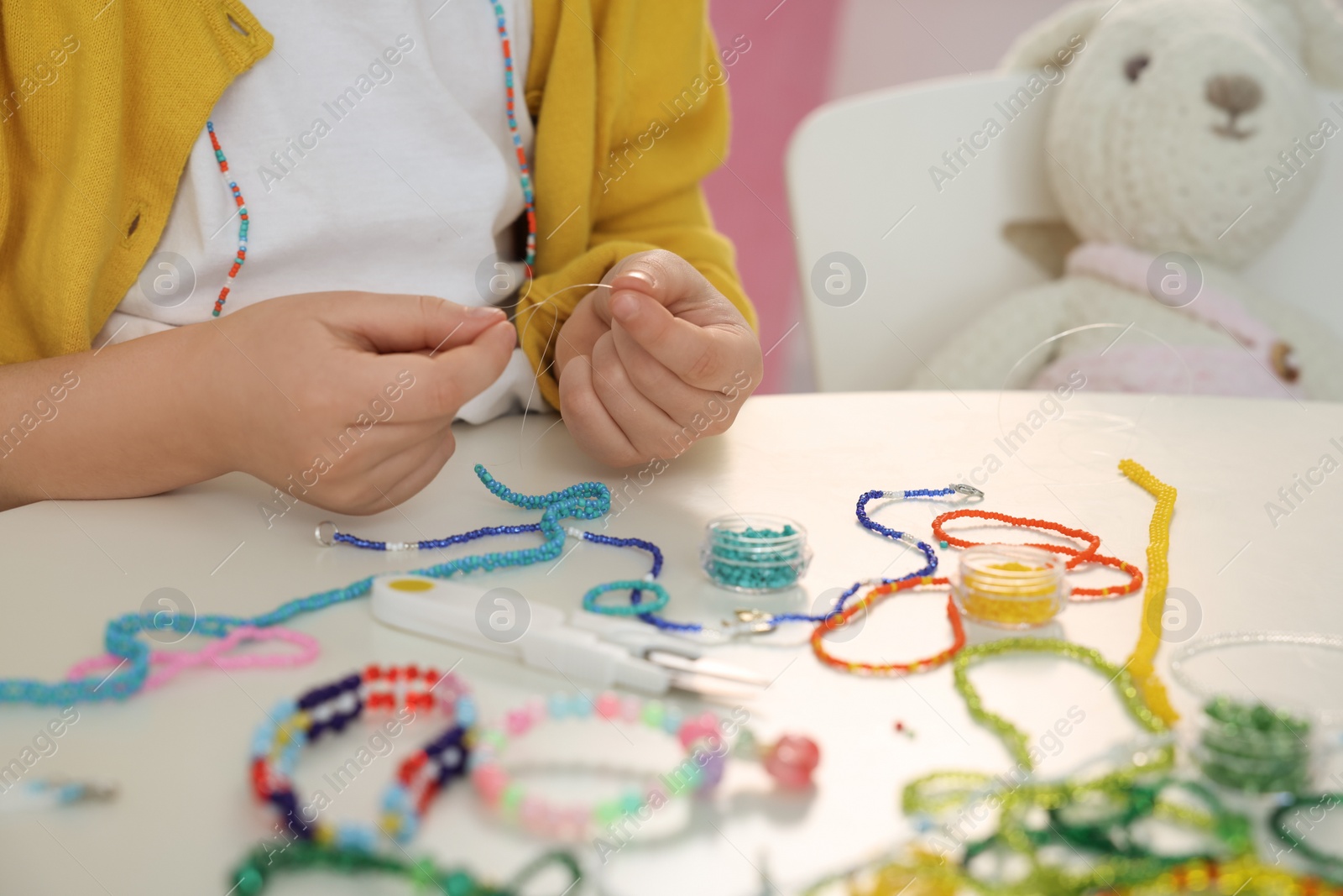 Photo of Little girl making beaded jewelry at table in room, closeup