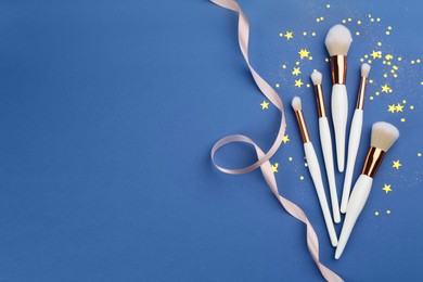 Different makeup brushes, ribbon and shiny confetti on blue background, flat lay. Space for text