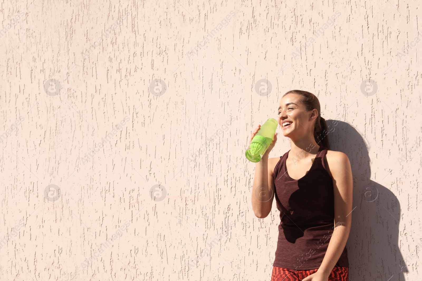 Photo of Young sporty woman drinking water from bottle near wall outdoors on sunny day. Space for text