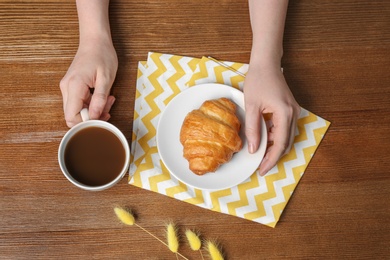 Photo of Young woman having breakfast with tasty croissant and cup of coffee at table, top view