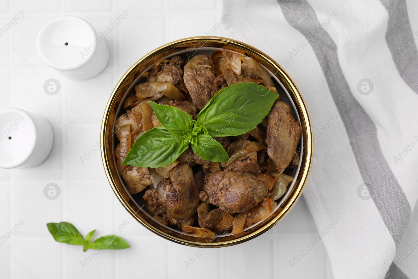 Photo of Delicious fried chicken liver with onion and basil in bowl on white tiled table, flat lay