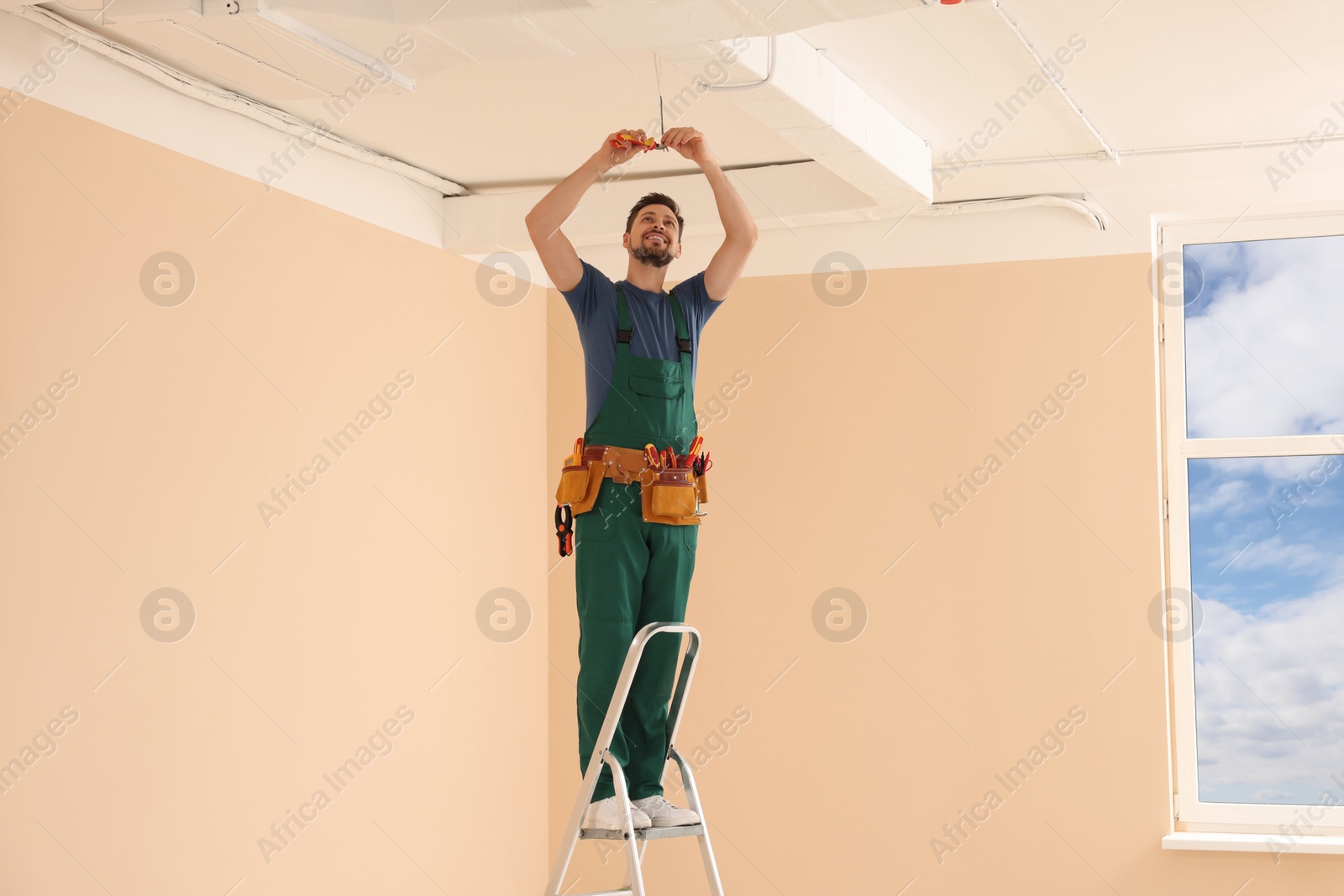 Photo of Electrician in uniform with pliers repairing ceiling wiring indoors