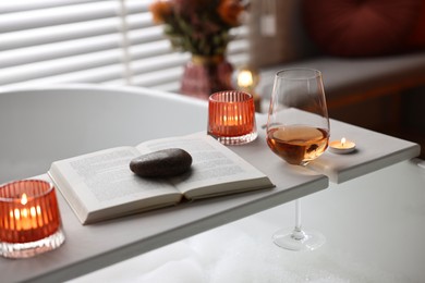 Photo of White wooden tray with glass of rose wine, book and burning candles on bathtub in bathroom