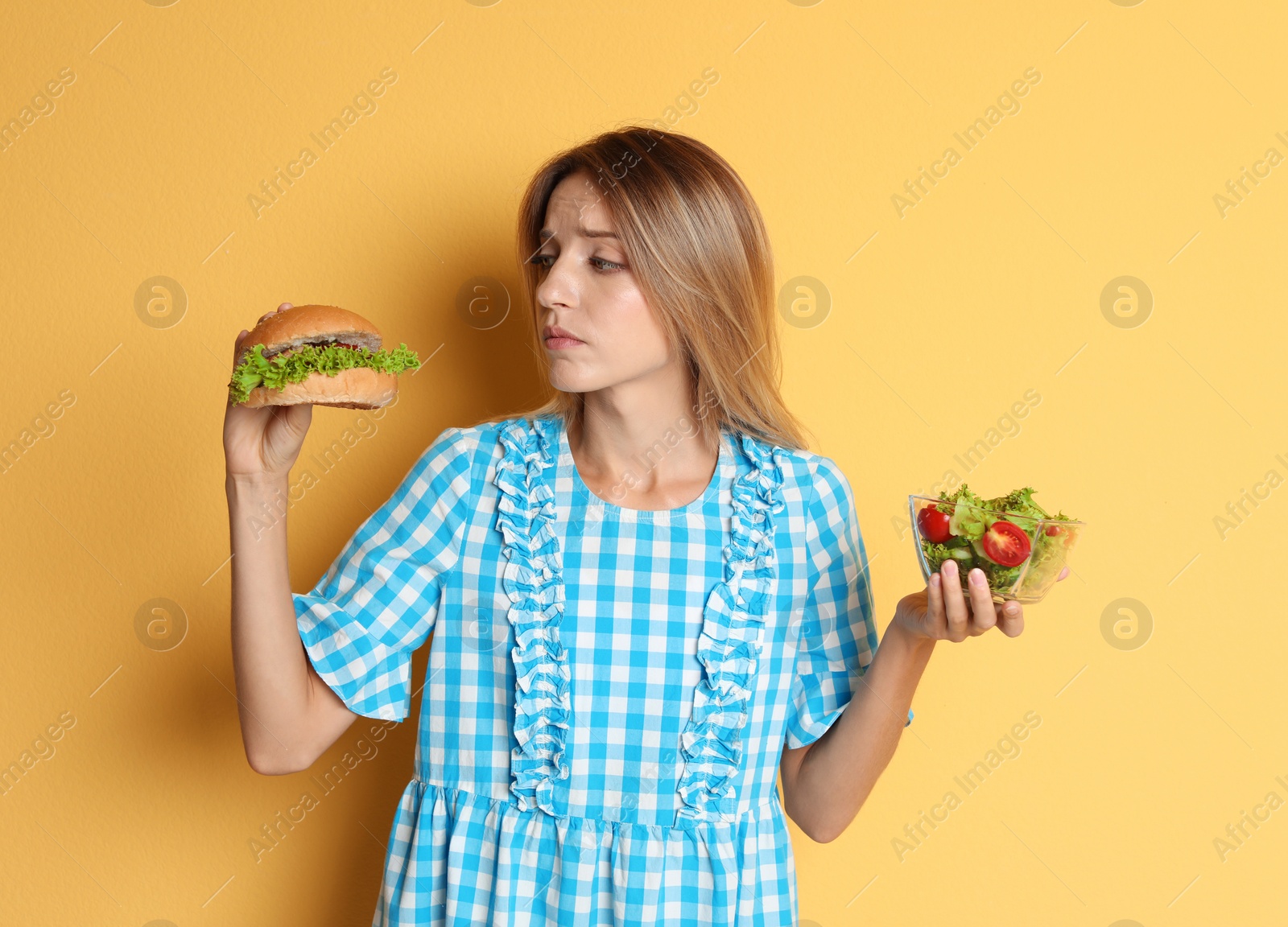 Photo of Young woman holding burger and salad on color background. Choice between diet and unhealthy food