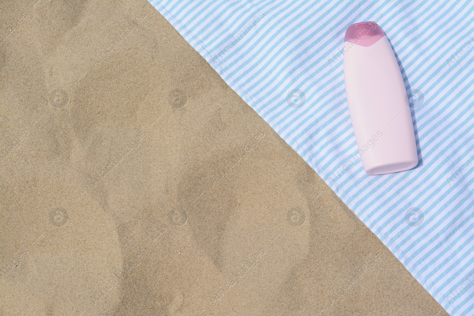 Photo of White and light blue striped towel with bottle of sunblock on sandy beach, top view. Space for text