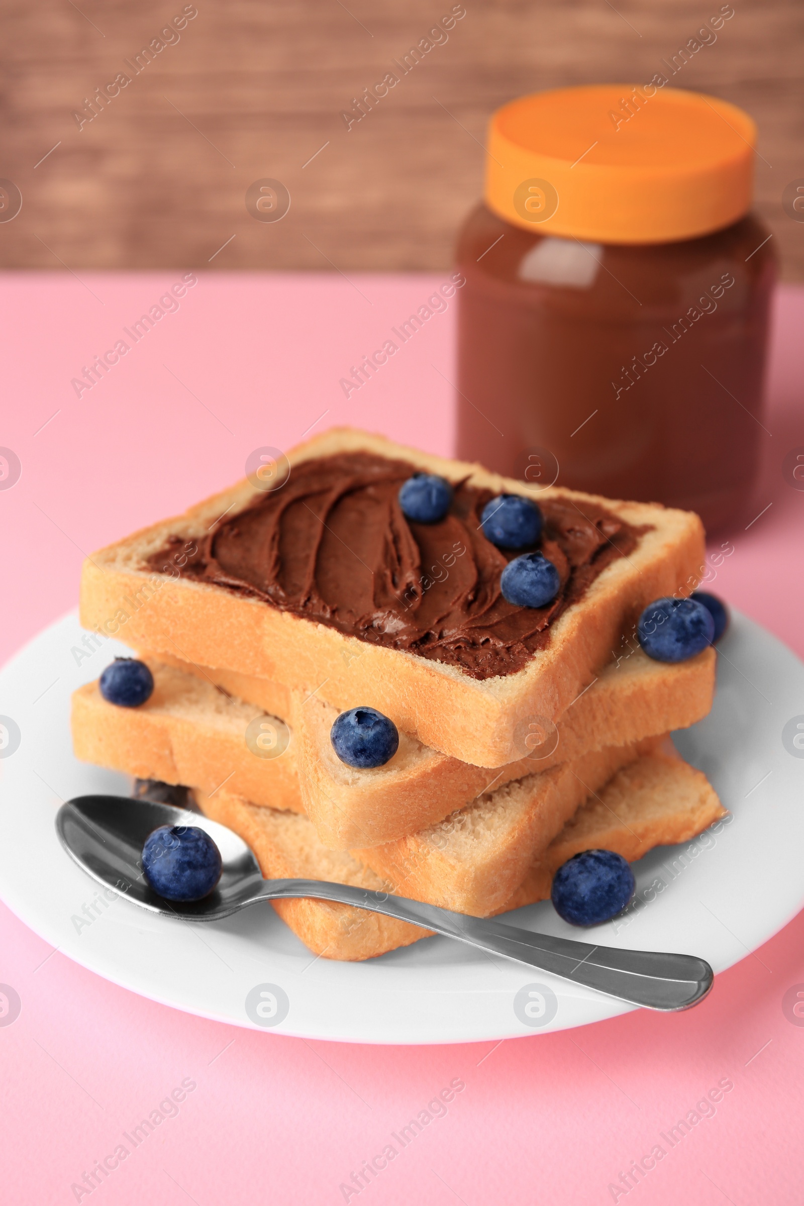 Photo of Tasty toast with chocolate paste and blueberries near jar on pink table