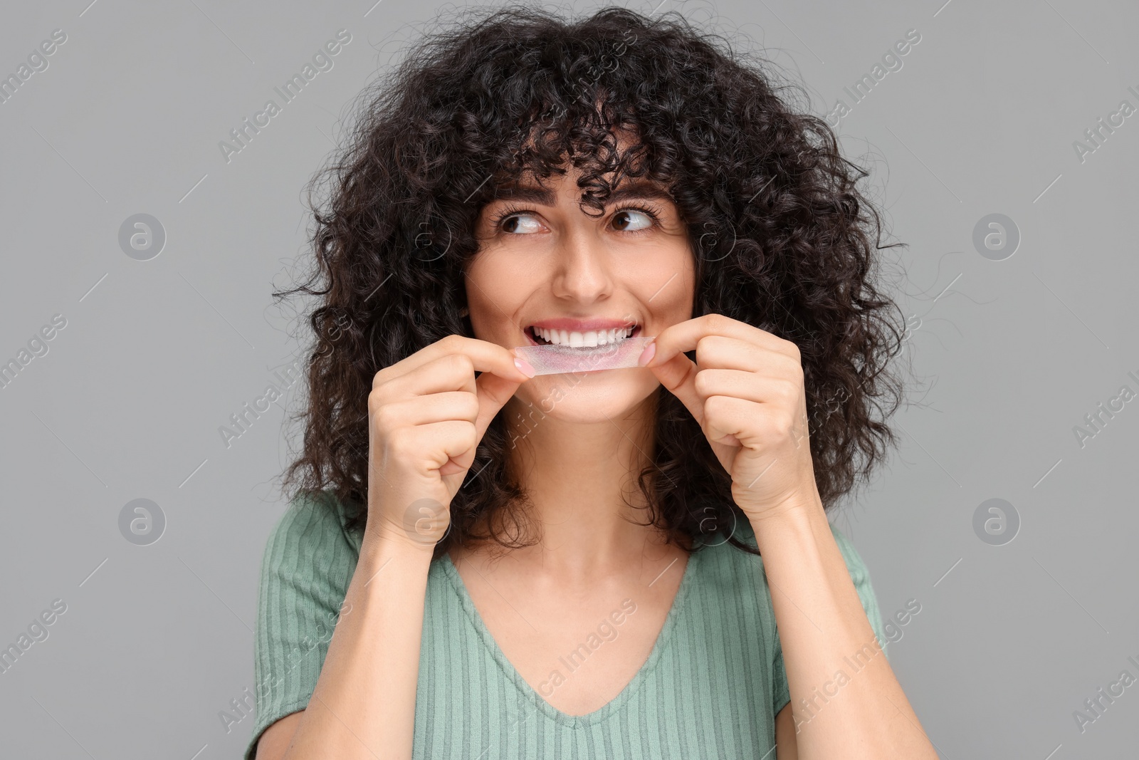 Photo of Young woman applying whitening strip on her teeth against grey background