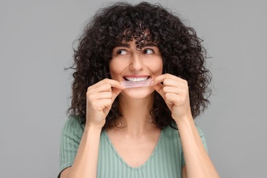 Young woman applying whitening strip on her teeth against grey background