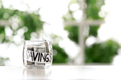 Photo of Glass jar with money and word SAVINGS on table against blurred background, space for text