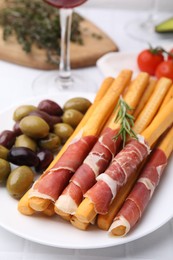 Photo of Plate of delicious grissini sticks with prosciutto and olives on white table, closeup