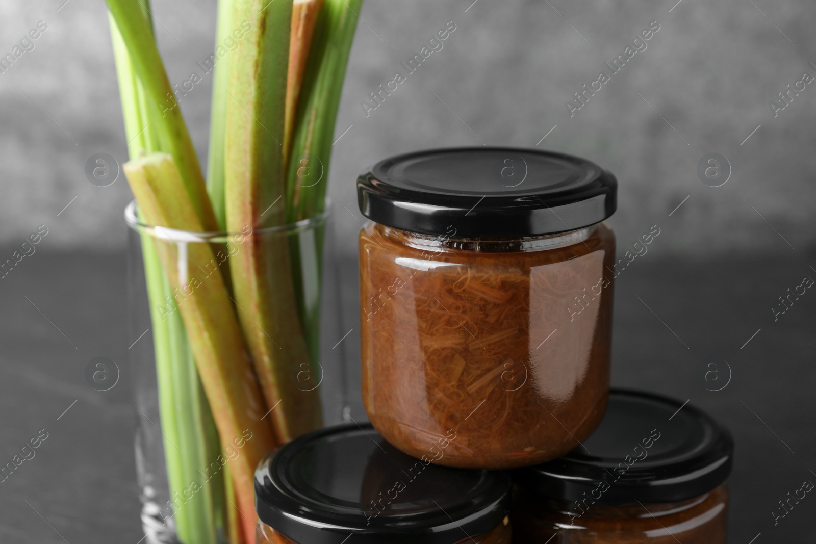 Photo of Jars of tasty rhubarb jam and stalks on grey table, closeup