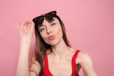 Photo of Beautiful young woman with sunglasses blowing kiss while taking selfie on pink background